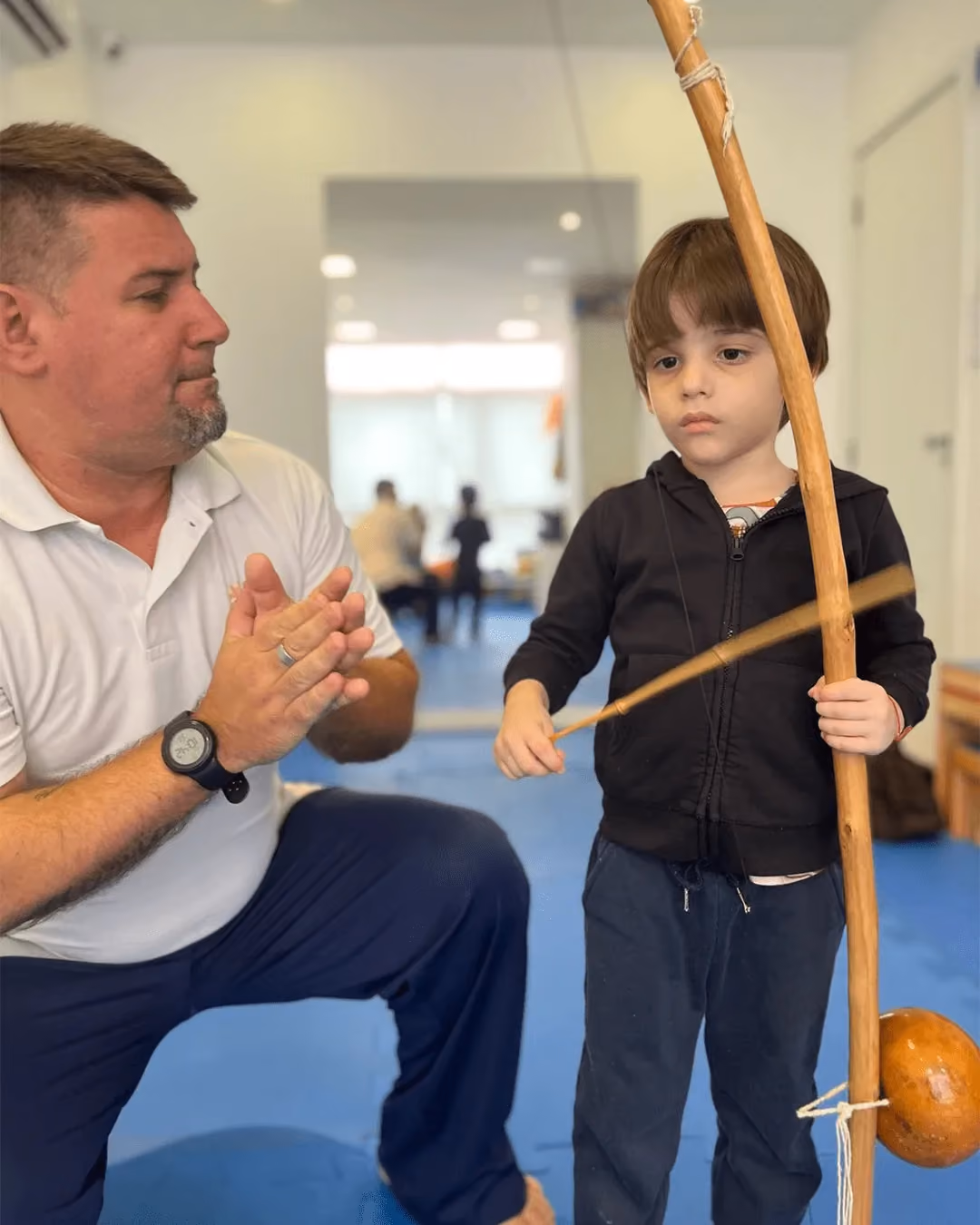 Capoeira teacher crouching and clapping beside a child holding a berimbau.