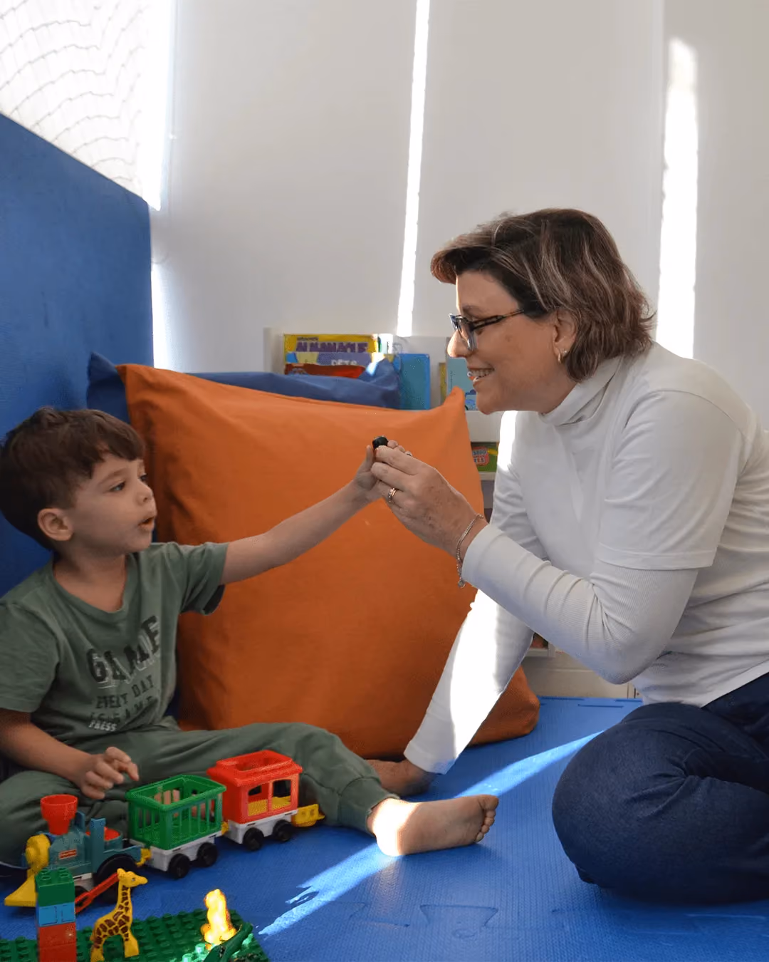 Fonoaudióloga e criança sentadas no tatame, brincando com um trem de brinquedo.
