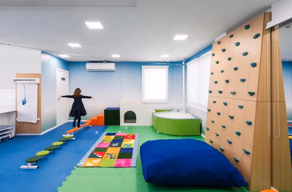 Sala de integração sensorial com parede de escalada, tapetes coloridos e circuito no chão.