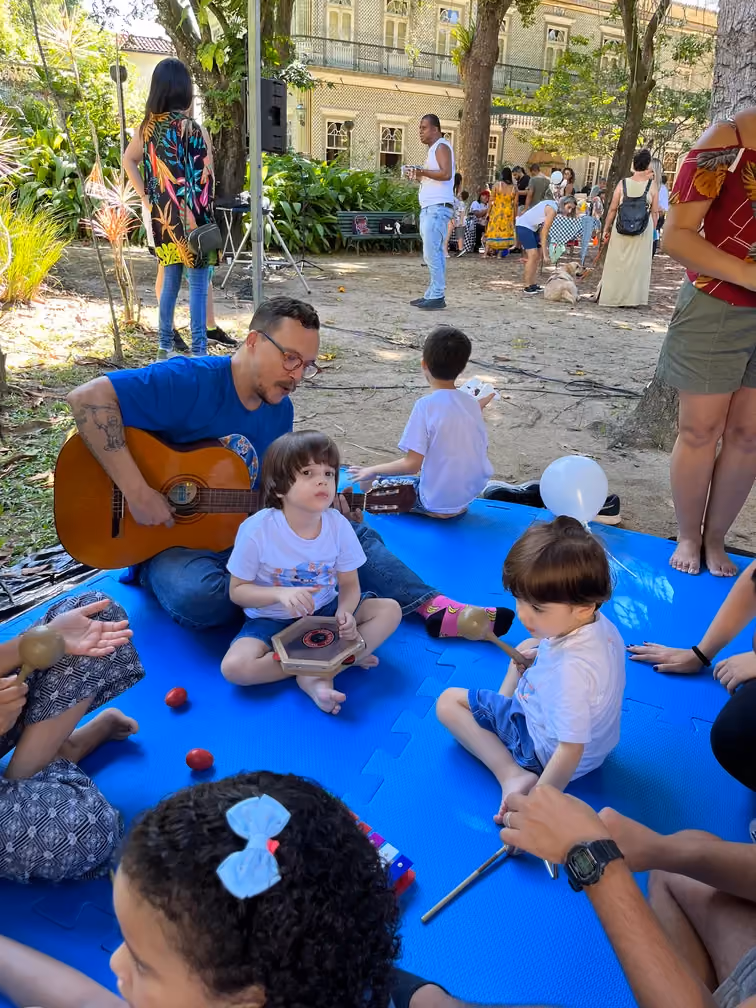 Roda de música ao ar livre, com terapeuta ao violão e crianças sentadas no tatame. - 4 de 4