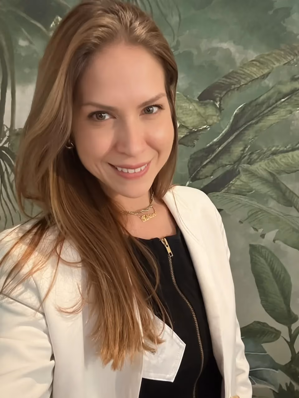 Dr. Tathiana de Almeida Martins de Souza smiling in a white blazer, in front of a leafy botanical wall